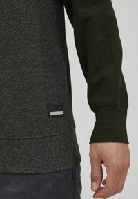 Donkergroene sweatshirt van een gestructureerde stof, met ribgebreide manchetten en een geweven label op de onderlinkerzijde met de tekst "ISOLID" in reliëf.