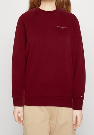 Osoba ubrana w bordową bluzę Tommy Hilfiger i beżowe spodnie, stojąca na jednolitym jasnym tle.