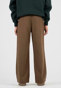 Bruine wijdpijpenbroek gemaakt van een zachte stof, met een gladde textuur en twee knopen aan de achterkant. Het model draagt een donkergroene top.