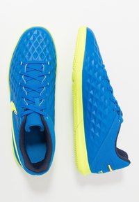 Zapatillas de fútbol sala en azul y amarillo con upper texturizado, cordones y cuellos acolchados, mostradas desde vistas superior y lateral sobre un fondo blanco.