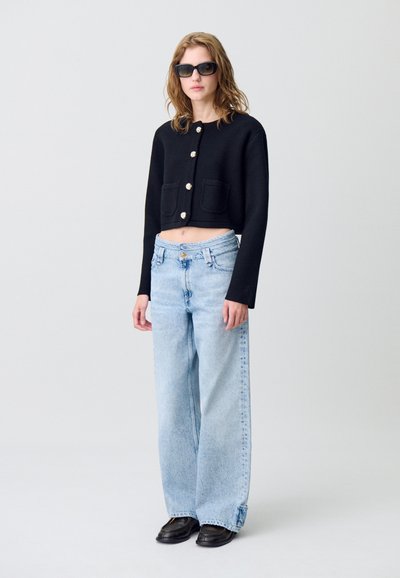 Cardigan nero corto con grandi bottoni e tasche, abbinato a jeans a palazzo blu chiaro e scarpe nere. Modello con occhiali da sole.