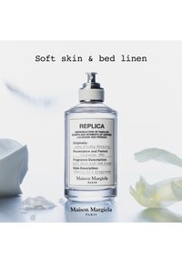 Gjennomsiktig flaske med Maison Margiela Replica parfyme merket Soft Skin og Bed Linen, plassert på en reflekterende overflate med mykt stoff og kronblader i nærheten.