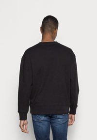 Uomo con capelli corti che indossa un felpa nera e jeans blu, in piedi con la schiena rivolta verso la fotocamera su uno sfondo bianco semplice.