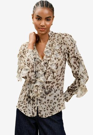 Transparente Bluse mit V-Ausschnitt, Rüschenelementen an Vorderseite und Ärmeln, in einem beigen und braunen Tierprintmuster, kombiniert mit dunklen Jeans.