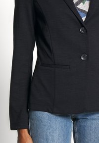 Veste noire avec une finition texturée, deux boutons et des poches latérales, associée à un jean bleu. Coupe ajustée et design taillé sur mesure.