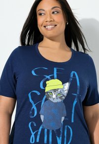 Tummansininen pehmeästä kankaasta valmistettu t-paita, jossa on kuvio kissasta, joka pitää keltaista hattua ja sinistä kuvioitua paitaa.