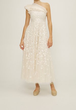 Vrouw die een mouwloze, een-schouder, witte kanten jurk tot aan de enkels draagt met bloemrijke borduursels en beige espadrille sleehakken.