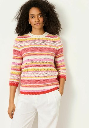 Frau mit lockigem Haar, die weiße Hose und einen mehrfarbig gestreiften Strickpullover mit 3/4-Ärmeln trägt, steht vor einem einfarbigen Hintergrund.