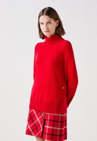 Czerwony sweter z wysokim prążkowanym kołnierzem, zdobiony guzikami po bokach oraz prążkowanym dołem. Zestawiony z czerwoną spódnicą w kratę z białymi liniami.