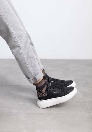 Black vegan leather sneakers with a leopard print accent at the heel, white rubber sole, and paired with gray rolled-up trousers and sparkly socks.