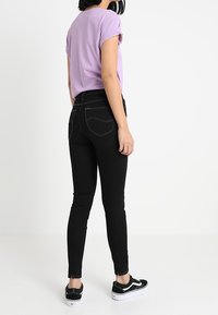 Jeans skinny negros con dos bolsillos traseros y costuras blancas, combinados con una camiseta lila y zapatillas negras. El corte es ajustado en las piernas.