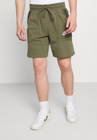 Muž v olivově zelených šortkách Nike Air, s bílými ponožkami a bílými teniskami se zelenými akcenty, stojí proti jednobarevnému pozadí.