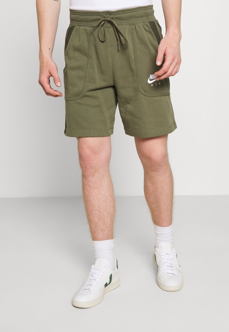 Muž v olivově zelených šortkách Nike Air, s bílými ponožkami a bílými teniskami se zelenými akcenty, stojí proti jednobarevnému pozadí.