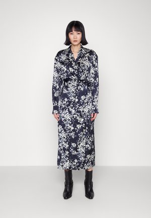 Kvinne står mot en enkel bakgrunn iført en langermet, svart og hvit blomstret midikjole med svarte ankelstøvler.