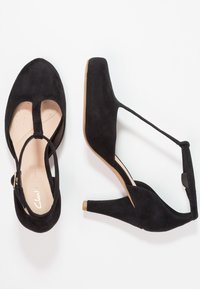 Mustat mokkanahkakengät korkokengät suljetulla kärjellä, t-nauhatyylillä, beige sisäpohja ja pieni metallikoriste korkolapasessa.