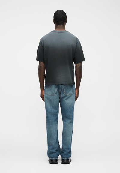 Άνδρας που στέκεται με την πλάτη στην κάμερα, φορώντας σκούρο μπλουζάκι με διαβάθμιση, μπλε τζιν και μαύρα παπούτσια, μπροστά σε απλό λευκό φόντο.