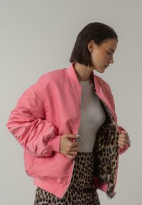 Rosa bomberjacka med en glänsande yta, ribbad krage och manschetter, med fodring i leopardmönster. Bärs öppen över en ljusgrå topp.