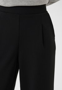 Gros plan sur un pantalon noir à taille haute avec des plis sur le devant, porté avec un pull côtelé gris clair rentré à l’intérieur.