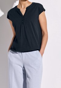 Svart kortärmad topp med V-ringning och fem knappar, i kombination med ljusgrå vida byxor. Mjuk tyg och avslappnad passform.