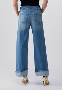 Inimene, kellel on jalas laiade säärtega sinised kägistatud teksad ja kuldsed kõrgete kontsadega sandaalid, näidatud tagantpoolt tavalise tausta ees.