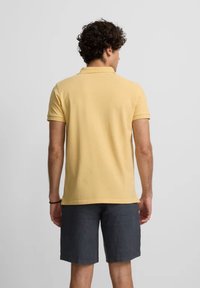 Camisa polo amarilla de manga corta con cuello estándar, combinada con pantalones cortos gris oscuro. Material de algodón con una textura suave.