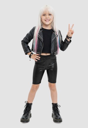Jeune fille aux longs cheveux blonds faisant le signe de la paix, portant une veste en cuir noire, un crop top, un short de biker et des bottes, souriant à la caméra.