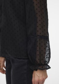 Blouse noire en tissu transparent à pois, manches longues et poignets à volants. Présente une bordure en dentelle à l'ourlet des manches. Motif texturé sur toute la surface.