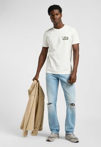 T-shirt bianca a maniche corte con logo verde "LEE", jeans strappati azzurri chiari e giacca beige appoggiata sul braccio, abbinati a sneakers grigie.
