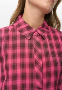 Vrouw in een roze en donkerbruin geruit overhemd met een puntige kraag, zichtbaar van nek tot borst.