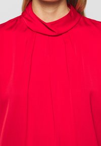 Blouse rouge à col bénitier haut, présentant des plis doux à l'avant et une texture lisse, fabriquée en tissu léger.