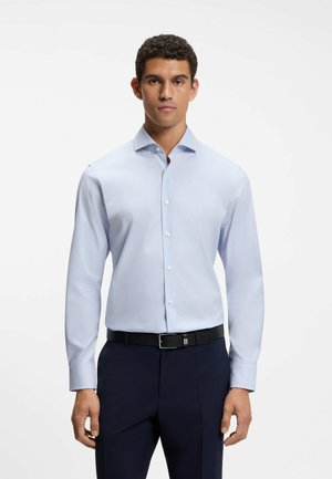 Mann mit hellblauem Hemd, schwarzem Gürtel und dunkelblauer Hose steht vor weißem Hintergrund.