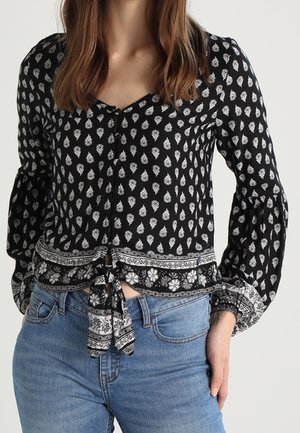 Vrouw met een zwarte blouse met lange mouwen, witte bloemenpatronen en een strik aan de voorkant, gecombineerd met lichtblauwe spijkerbroek.