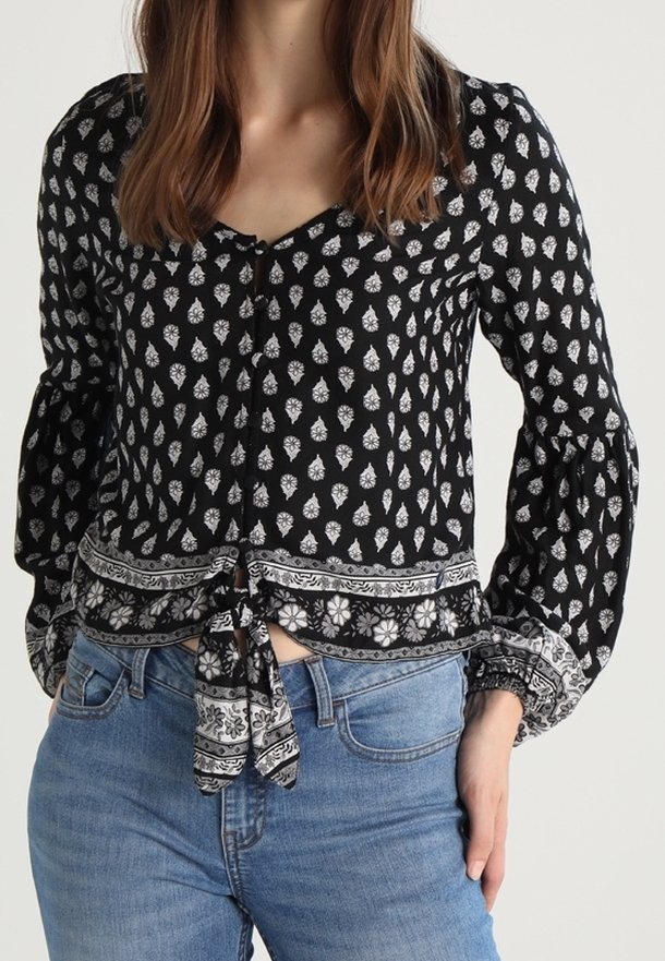 Femme portant une blouse noire à manches longues avec des motifs floraux blancs et un nœud à l'avant, associée à un jean bleu clair.