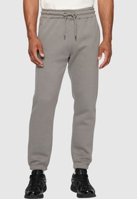 Grå joggebukser med elastisk midje, snøring og mansjetter ved anklene. Glatt tekstur, laget av mykt stoff. Svarte sneakers brukt.