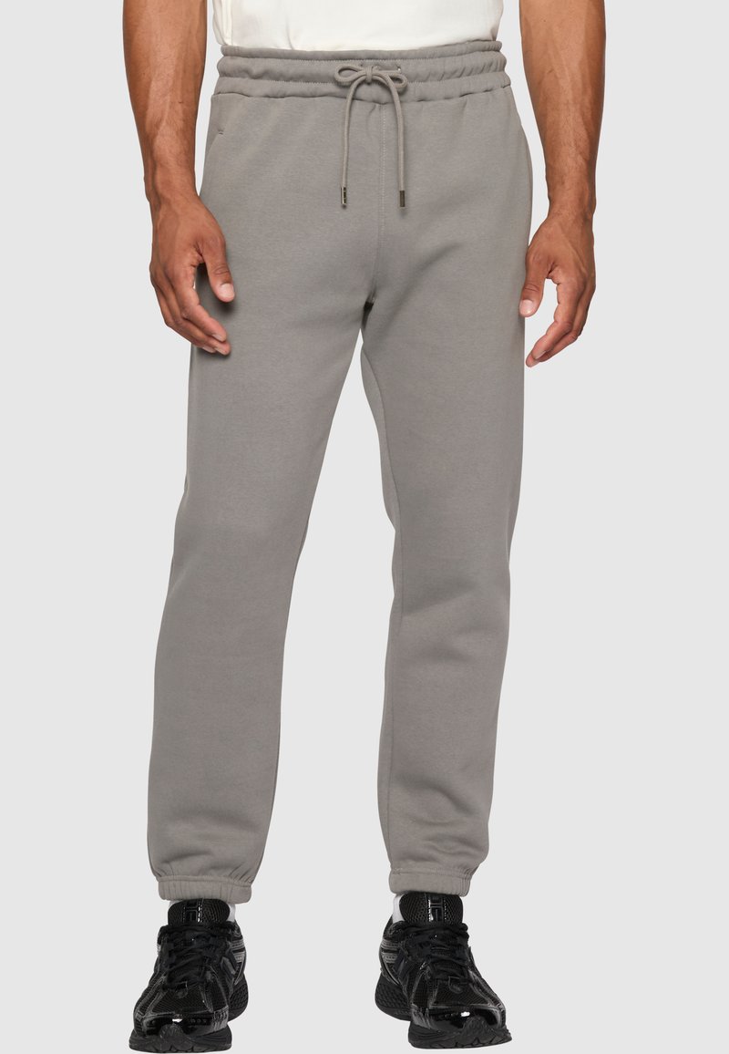 Grå joggebukser med elastisk midje, snøring og mansjetter ved anklene. Glatt tekstur, laget av mykt stoff. Svarte sneakers brukt.