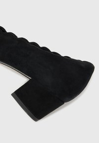 Black suede high-heeled shoe with a chunky heel. Features scalloped upper edge and a smooth texture. Rounded toe design, minimal accents.