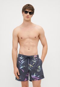 Čierne plavky s farebnými kvetinovými vzormi, elastický pás, šnúrka na stiahnutie a bočné vrecká; nosené s okuliarmi a odhaleným torzom.