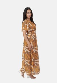 Vestido maxi en un patrón floral marrón y naranja, con cintura fruncida, mangas cortas y tejido fluido. Combinado con tacones transparentes.