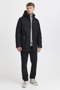 Svart vanntett jakke med hette, to frontlommer og hardware i kobberfarge. Bærer over en grå genser og svarte bukser, med grå sneakers.
