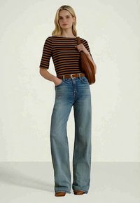 Blond kvinne iført navy- og oransje stripet topp, høy midje bred bukse, brun belte, brune sko, og bærende en brun skulderveske.