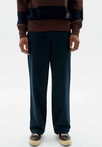 Dunkelblaue weit geschnittene Hosen aus weichem Stoff, kombiniert mit einem braun-blauen gestreiften Strickpullover und dunkelbraunen Schuhen mit beigen Sohlen.