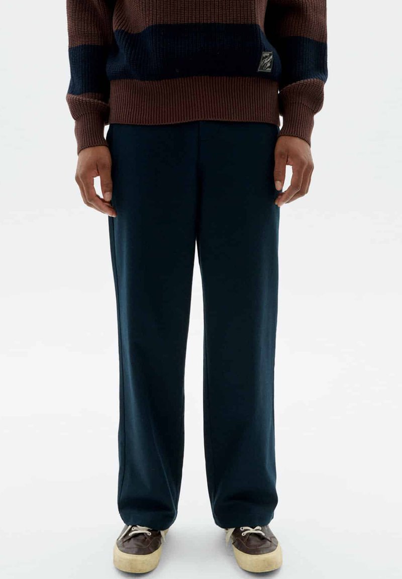 Dunkelblaue weit geschnittene Hosen aus weichem Stoff, kombiniert mit einem braun-blauen gestreiften Strickpullover und dunkelbraunen Schuhen mit beigen Sohlen.