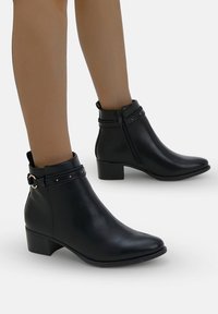 Stivali in pelle nera alti fino alla caviglia con tacco basso a blocco, cerniere laterali e cinturini decorativi con borchie in metallo e fibbie, indossati su gambe scoperte.