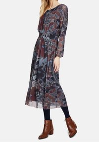 Lång blommig klänning med ett flerfärgat mönster i mörka nyanser, genomskinligt tyg, långa ärmar, samlad midja och ankellång fåll. Bruna ankelstövlar fulländar looken.