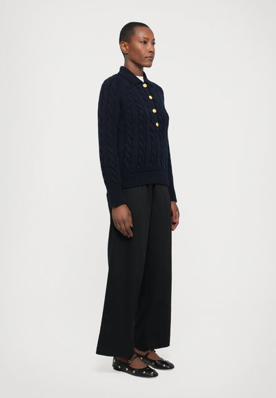 Pull en maille torsadée bleu marine avec un col et quatre boutons en or, associé à un pantalon large noir et des ballerines noires.