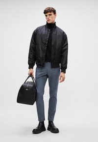 Schwarze Satin-Bomberjacke über einem schwarzen Rollkragenpullover, kombiniert mit hellblauen Slim-Fit-Hosen und schwarzen Leder-Stiefeletten. Tragend eine schwarze Sporttasche.
