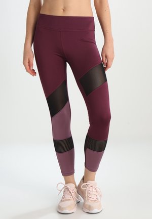 Kvinde iført bordeaux og lilla leggings med sorte meshpaneler, stående barfodet mod en ensfarvet lys baggrund.