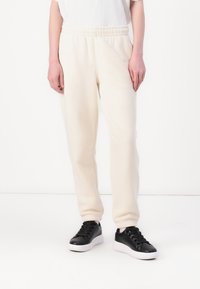 Ljusbeiga sweatpants med elastisk midja och sidofickor, kombinerat med svarta sneakers med vit sula. Slät textur.