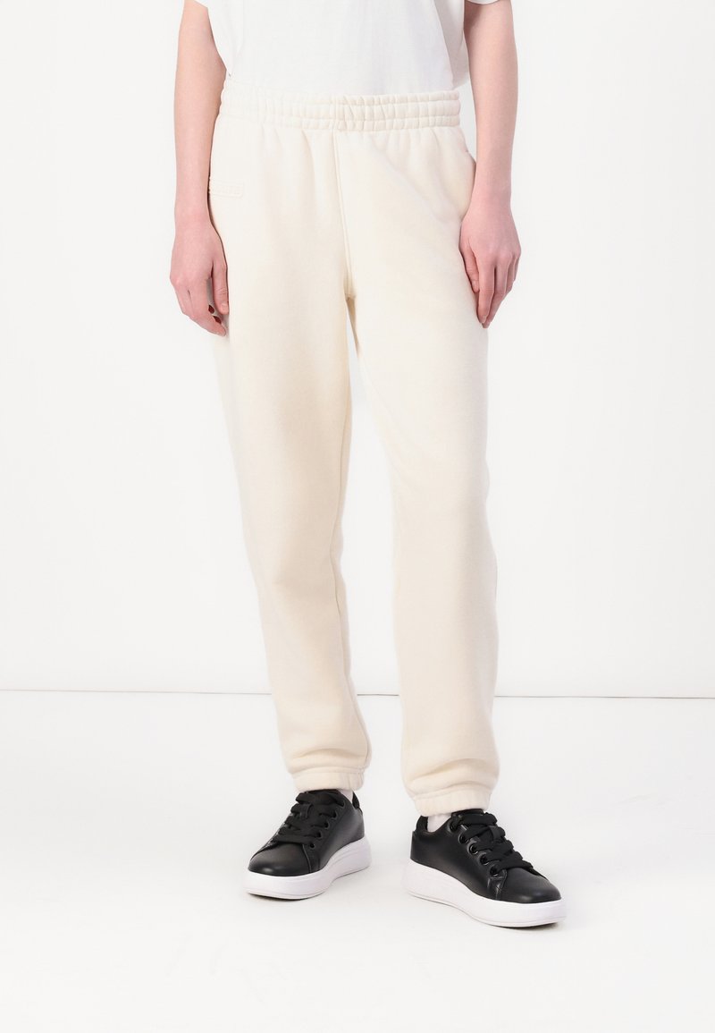 Ljusbeiga sweatpants med elastisk midja och sidofickor, kombinerat med svarta sneakers med vit sula. Slät textur.
