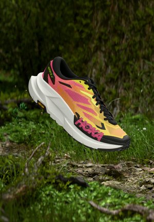 Scarpa da corsa con tomaia arancione e rosa, lacci neri e suola bianca spessa che fluttua sopra il terreno della foresta con fogliame verde e rocce.